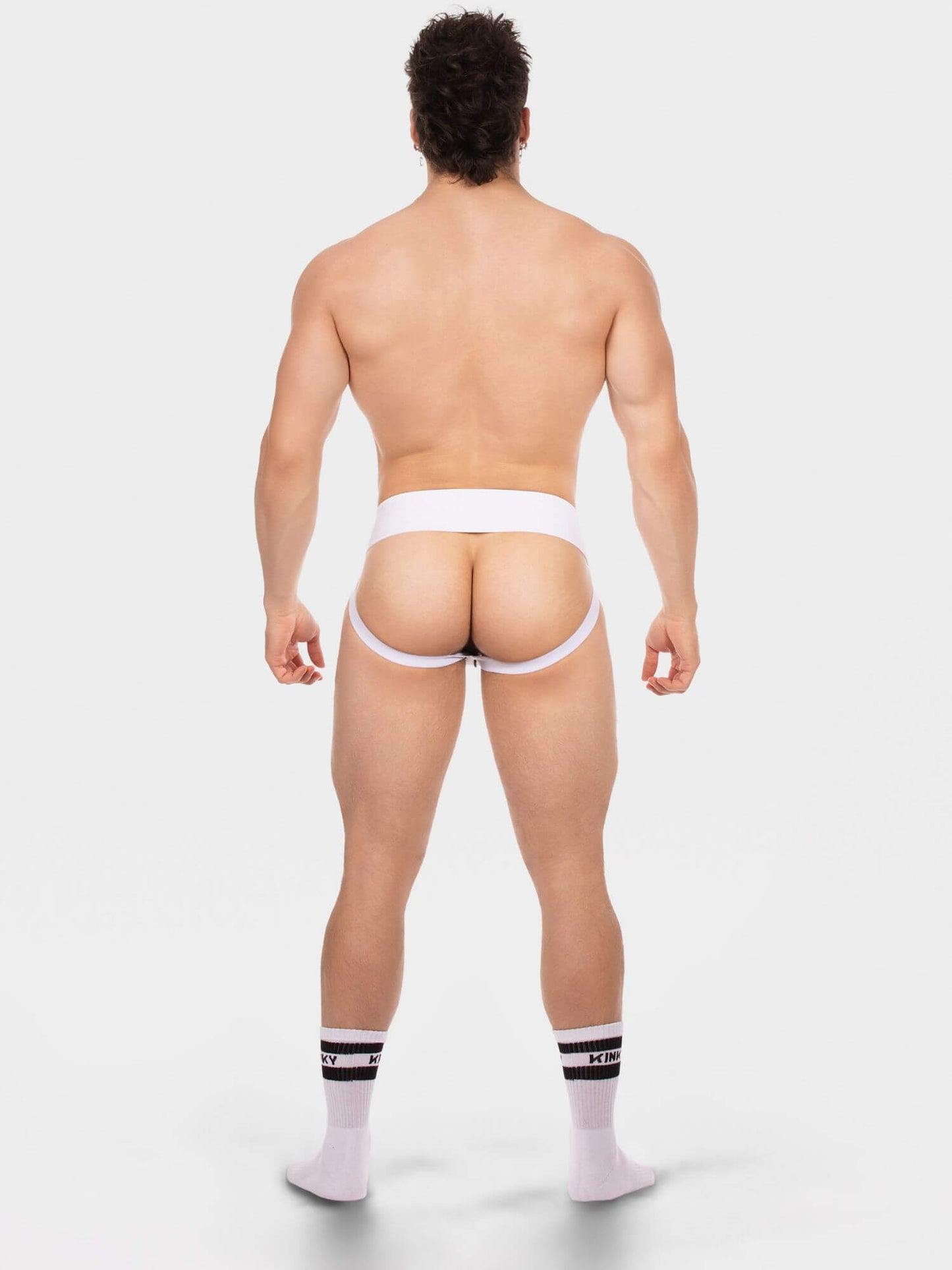Rear view of a muscular man wearing white men's jock and white crew socks with black stripes, standing against a light gray studio background.