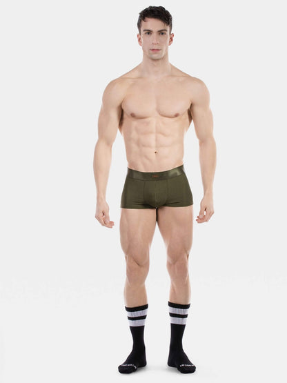 A shirtless male model stands against a light gray studio background wearing olive green boxer briefs and black crew socks with white stripes.