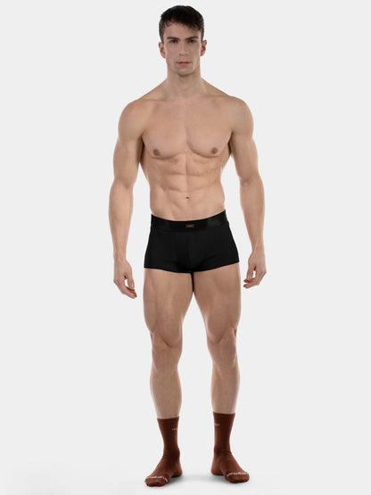 A muscular male model standing against a light gray studio background, wearing black boxer briefs and brown socks.