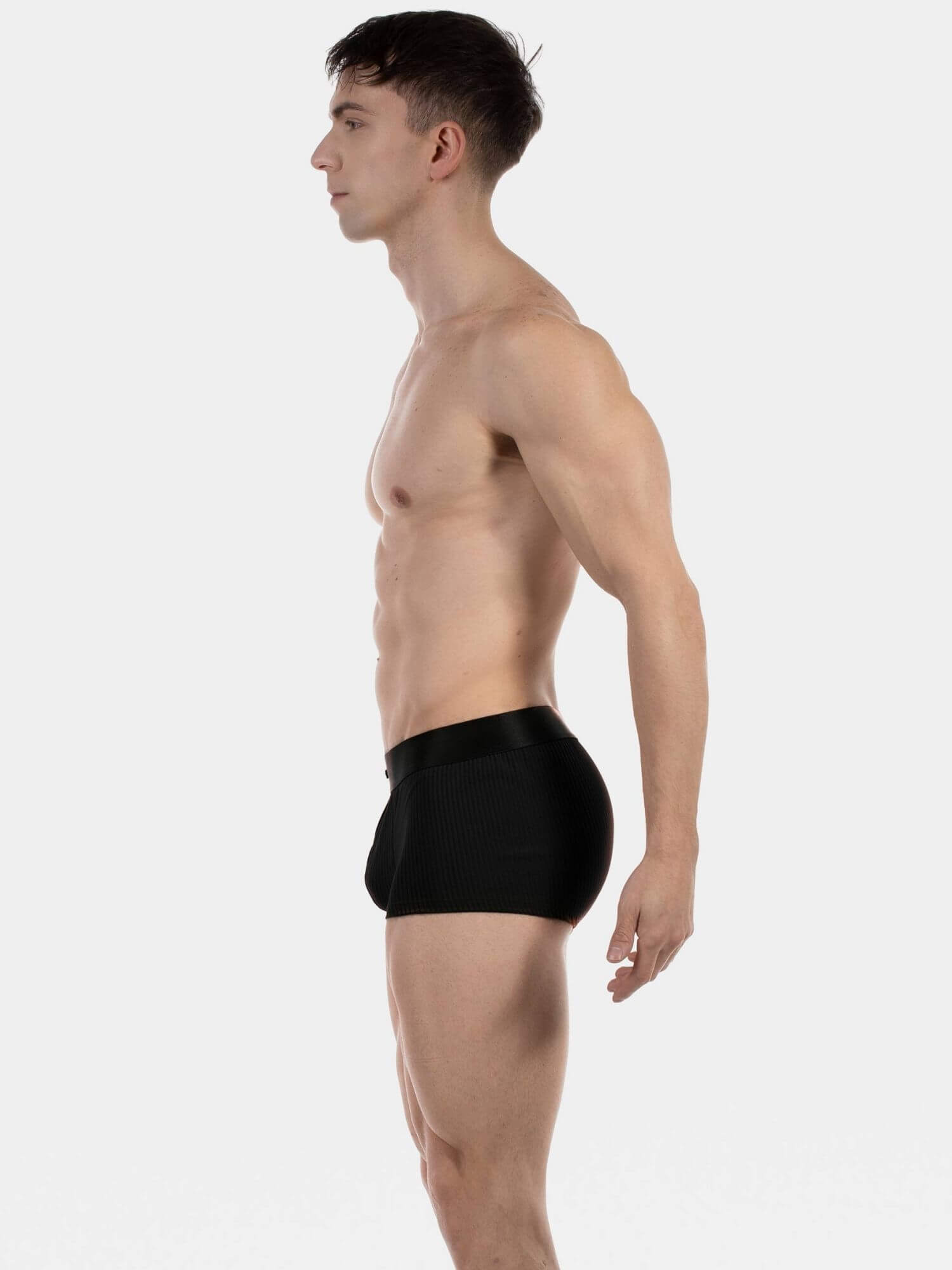 A man shown in side profile wearing black men's briefs against a plain light gray studio background.