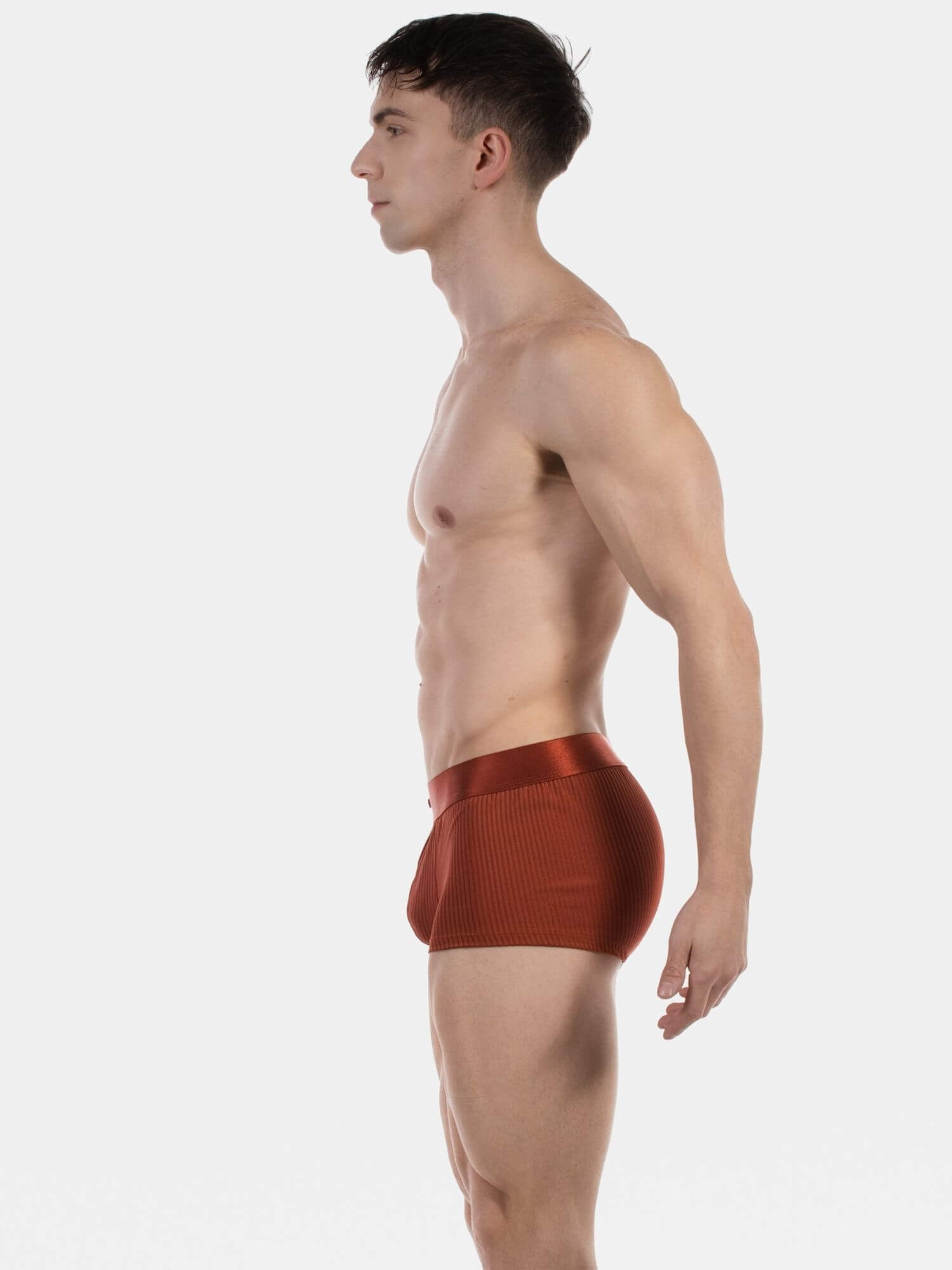 A man shown in side profile wearing textured, ribbed reddish-brown boxer briefs against a plain light gray studio background.