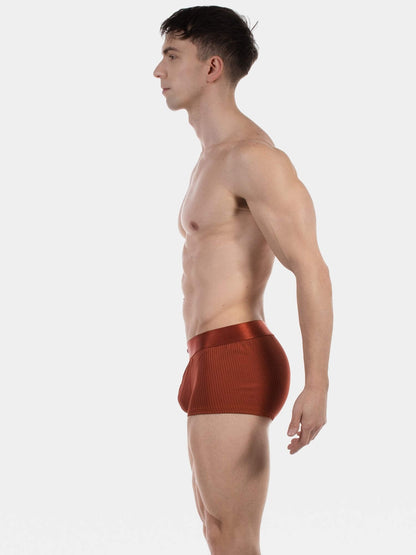A man shown in side profile wearing textured, ribbed reddish-brown boxer briefs against a plain light gray studio background.