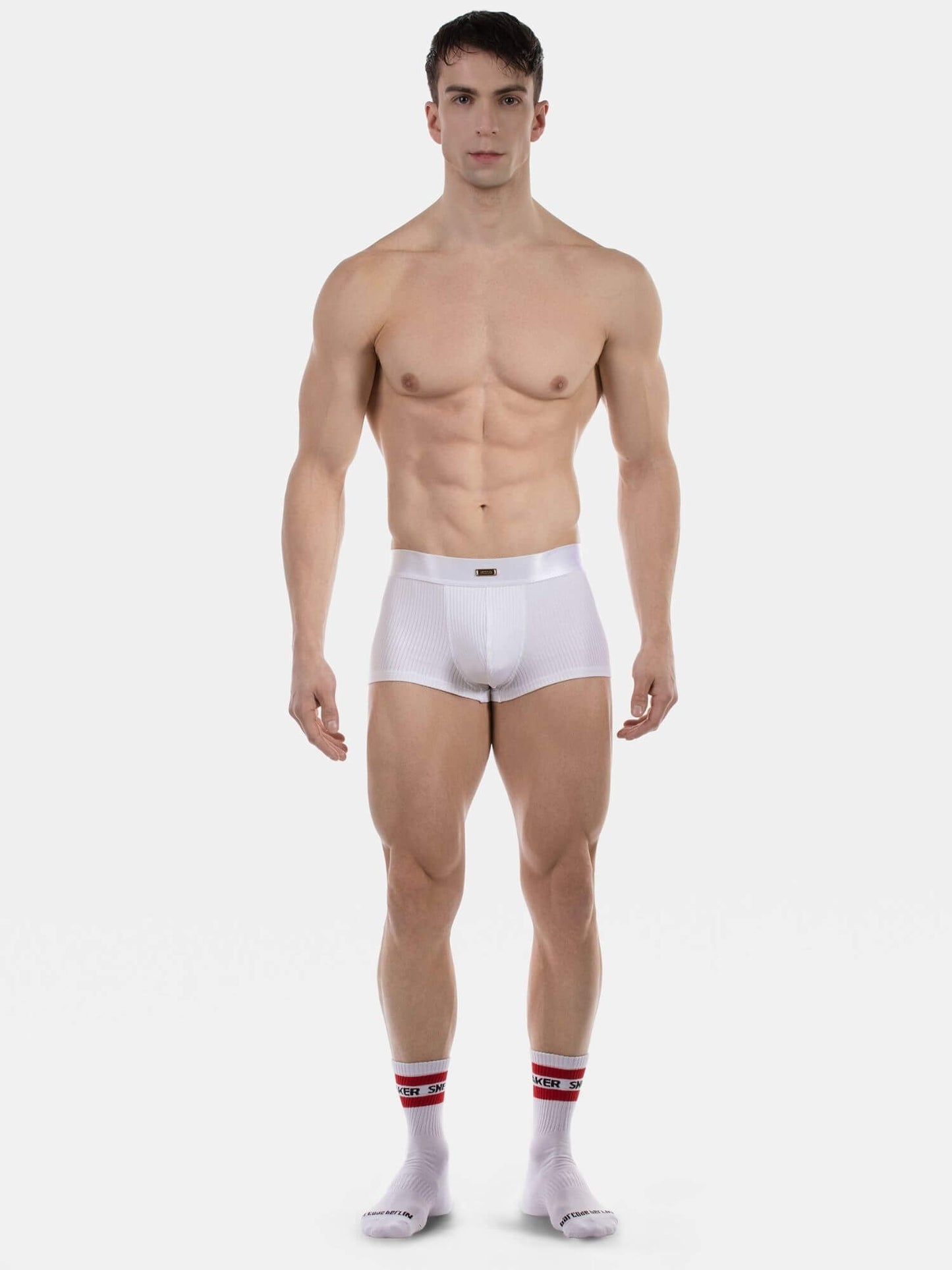 A muscular male model stands against a light gray studio background wearing white boxer briefs and white socks with red stripes.