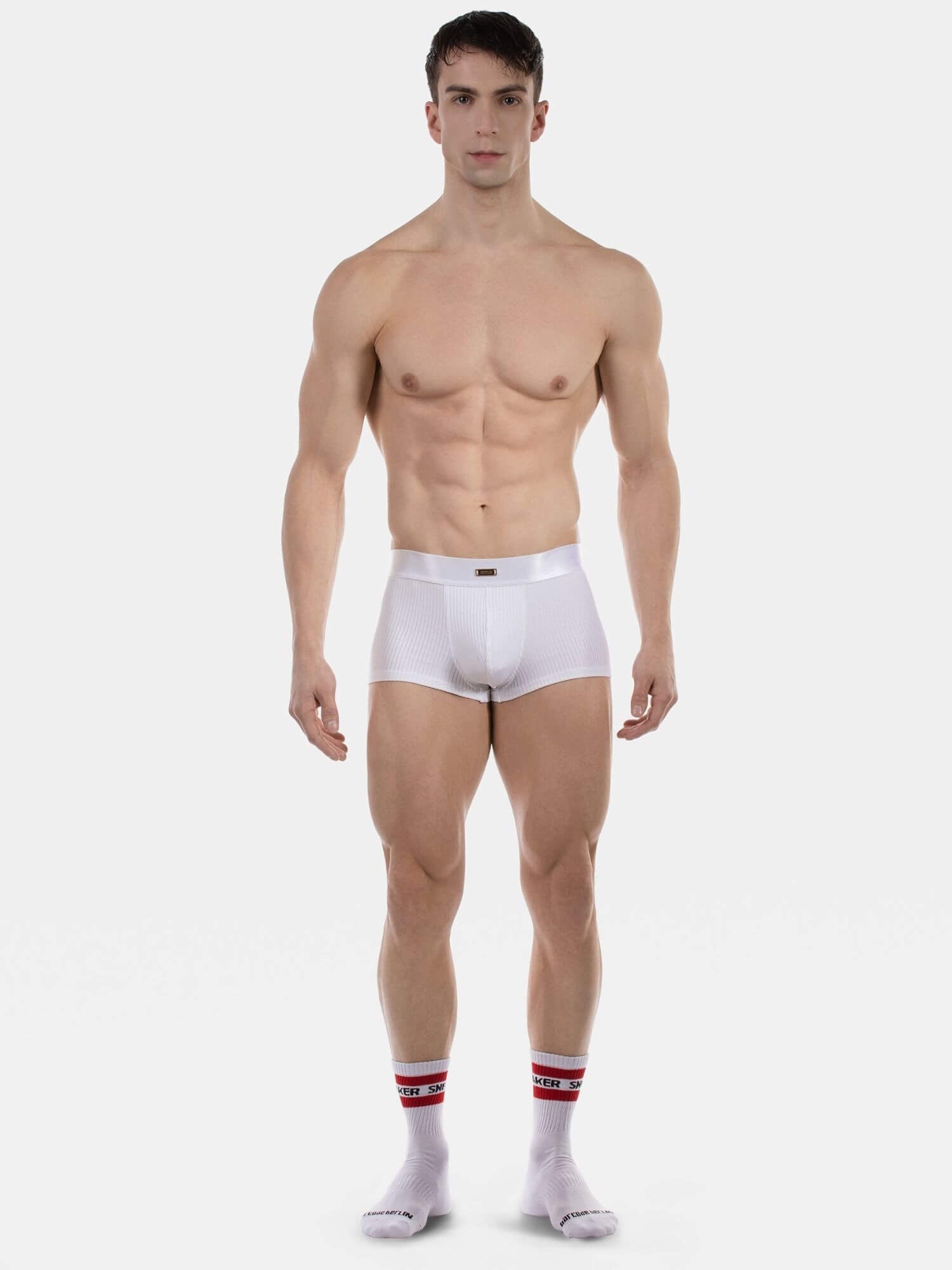 A muscular male model stands against a light gray studio background wearing white boxer briefs and white socks with red stripes.