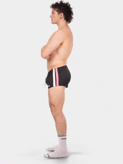 A side-profile studio photo of a shirtless man wearing black athletic shorts with vertical pink and white stripes, paired with light pink socks, against a plain light gray background.