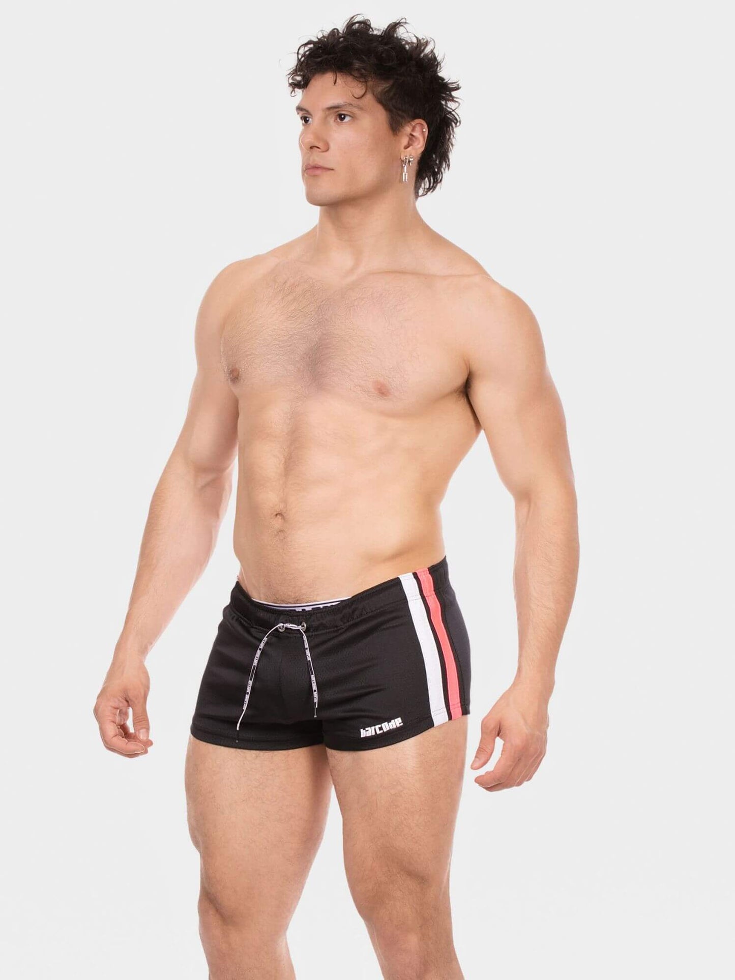 A shirtless man modeling black athletic swim trunks with white and pink side stripes and a white drawstring, photographed against a light gray studio background.