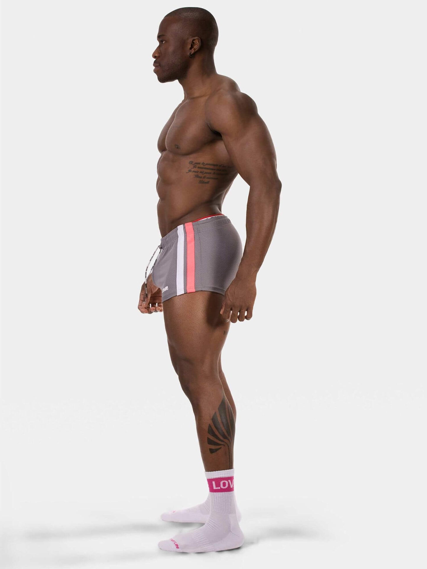 A muscular man in profile wearing gray athletic shorts with pink and white side stripes, paired with light pink socks, against a plain light gray studio background.