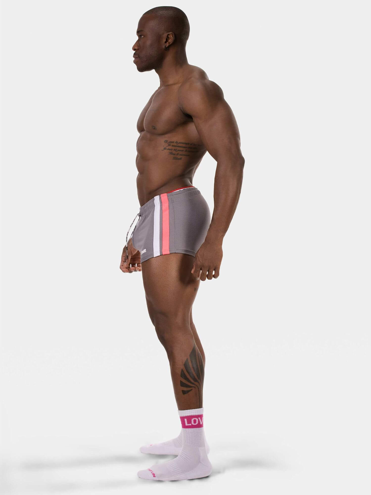 A muscular man in profile wearing gray athletic shorts with pink and white side stripes, paired with light pink socks, against a plain light gray studio background.
