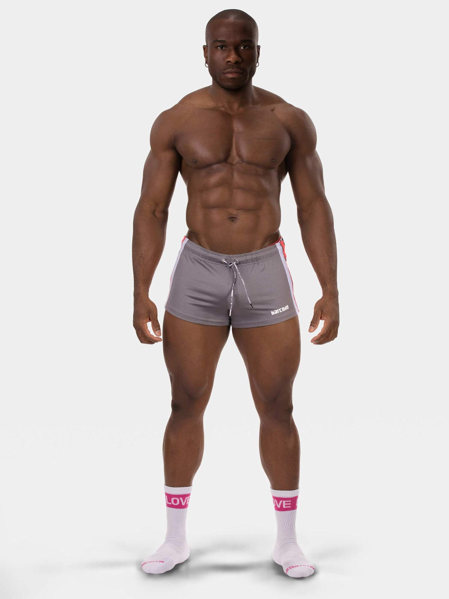 A muscular man modeling gray athletic shorts with a white drawstring and pink side stripes, paired with white socks, against a light gray studio background.