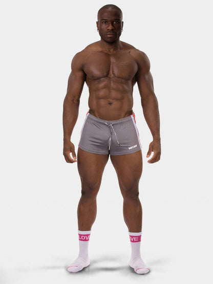 A muscular man modeling gray athletic shorts with a white drawstring and pink side stripes, paired with white socks, against a light gray studio background.