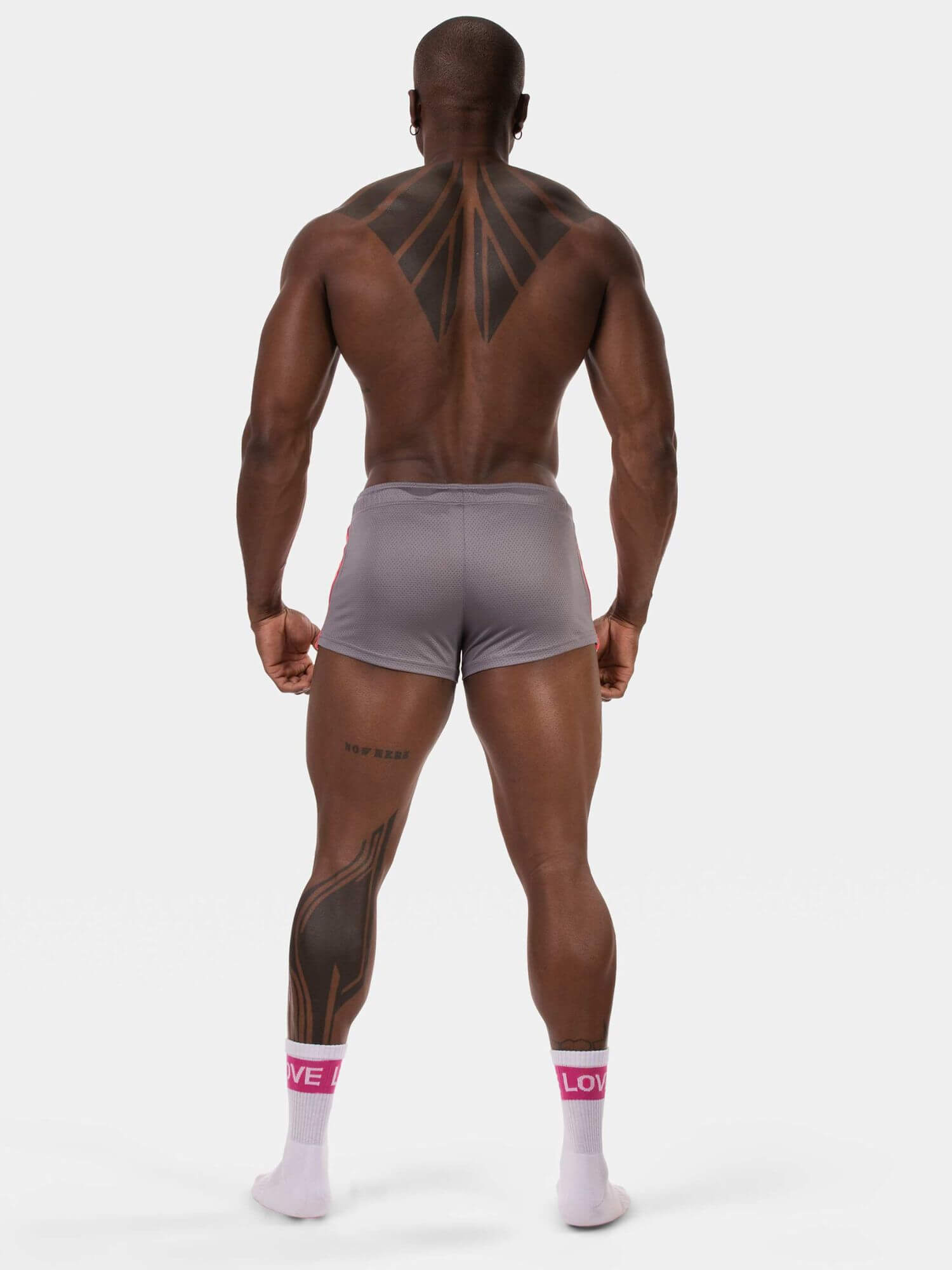 A muscular man viewed from the back wearing light gray athletic shorts and white crew socks with pink branding, standing against a plain light gray studio background.