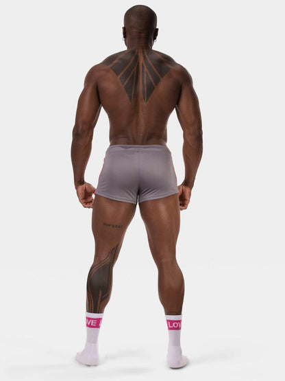 A muscular man viewed from the back wearing light gray athletic shorts and white crew socks with pink branding, standing against a plain light gray studio background.
