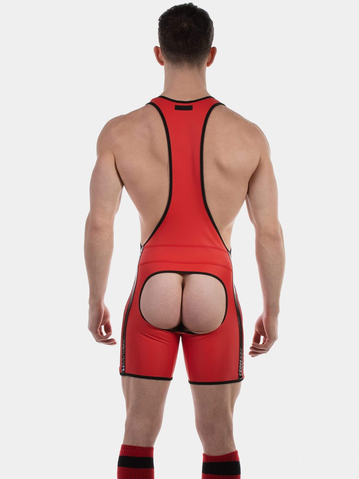 A muscular male model wearing a bright red wrestling singlet with black trim, paired with red and black socks, against a solid light gray background.