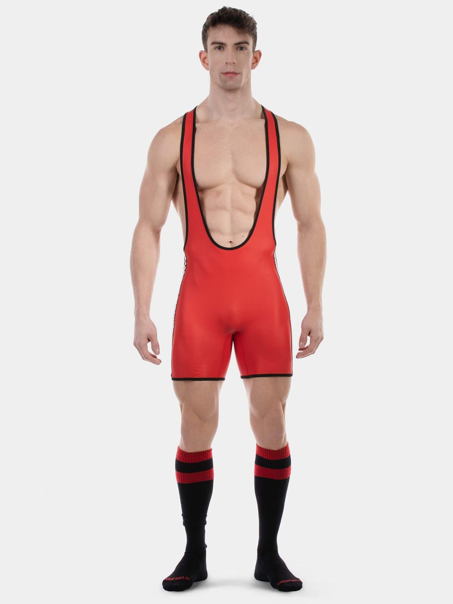 A muscular male model wearing a bright red singlet-style athletic romper with black trim, paired with black knee-high socks featuring red stripes, against a light gray studio background.