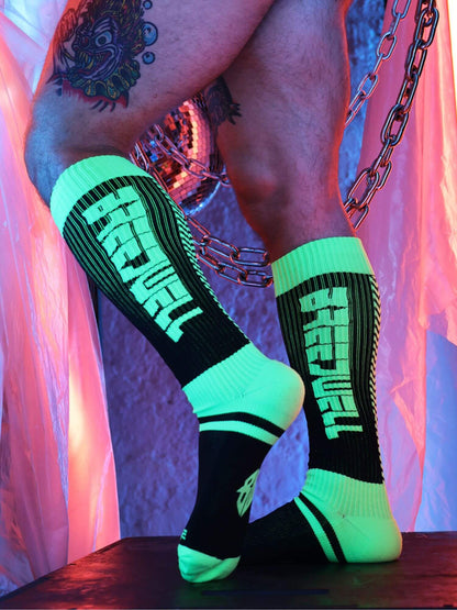 Person wearing black and green socks with branding, standing against a colorful background.