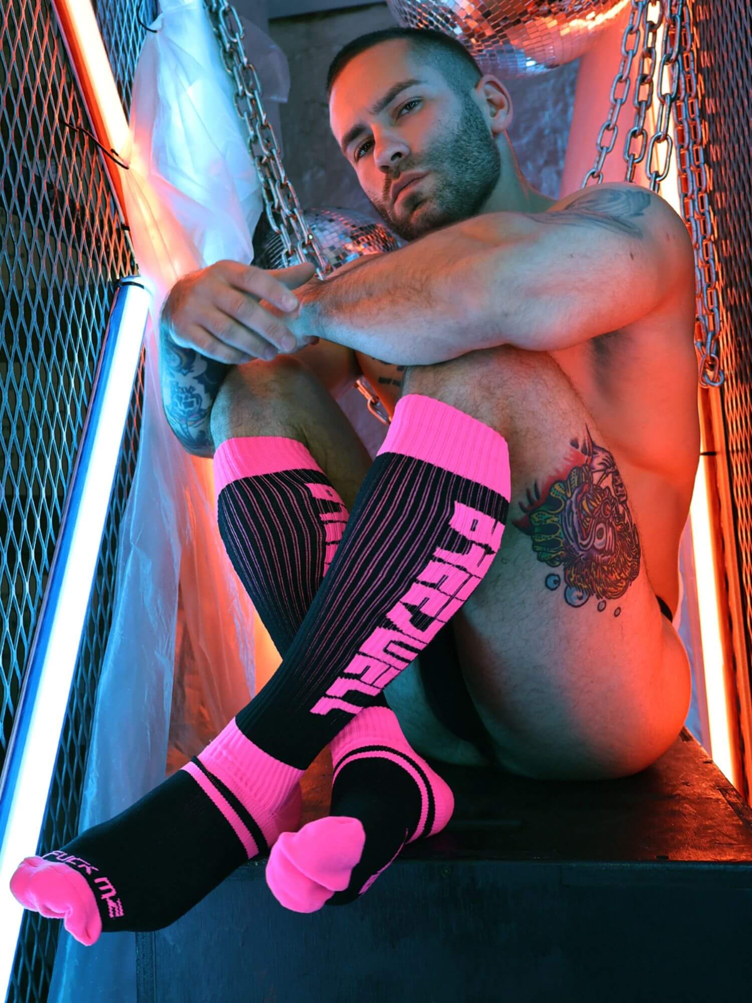 Man wearing black and pink socks with visible tattoos, sitting against a colorful background.