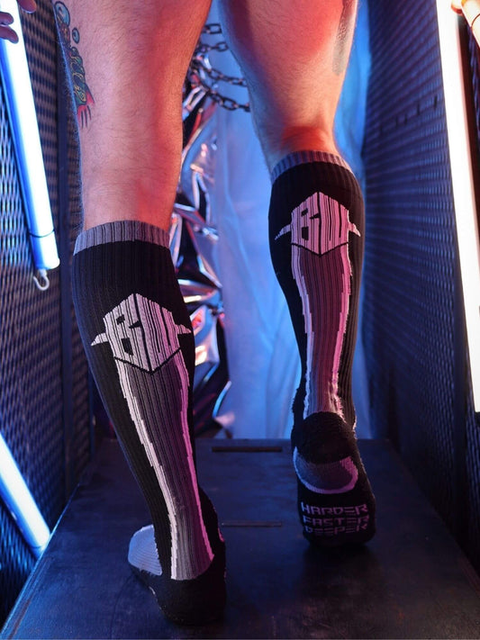 A person wearing black compression socks with gray and pink striped patterns and white logo graphics, standing on a dark platform in a neon-lit environment.