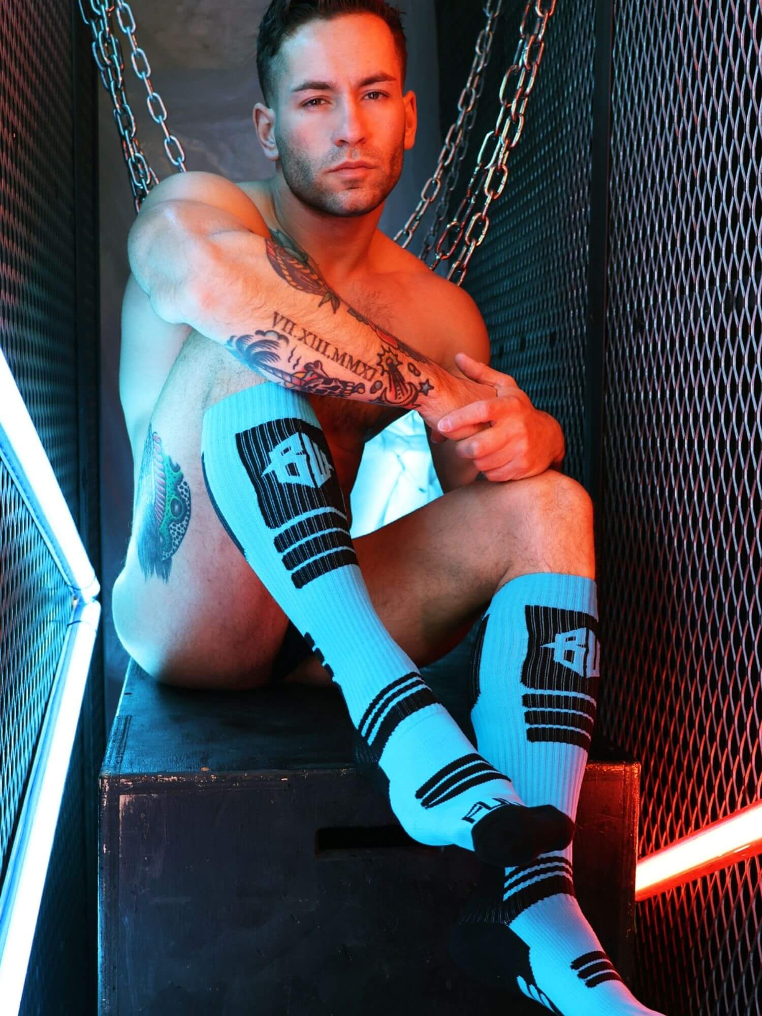 Man with tattoos wearing aqua socks with black designs sitting against a dark background.