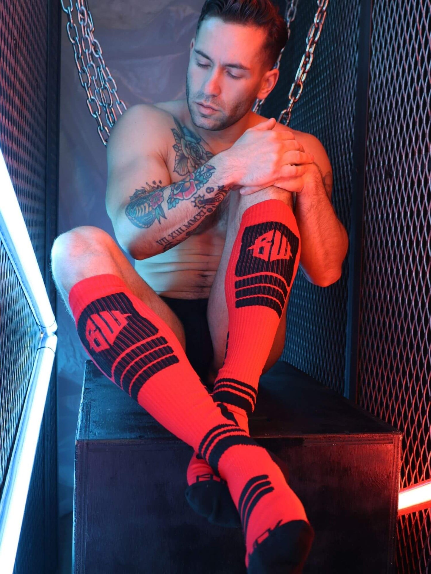 Man wearing red and black socks with visible branding, sitting in a dark room.
