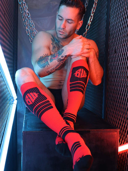 Man wearing red and black socks with visible branding, sitting in a dark room.