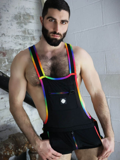 Man wearing a black outfit with rainbow suspenders against a textured wall.