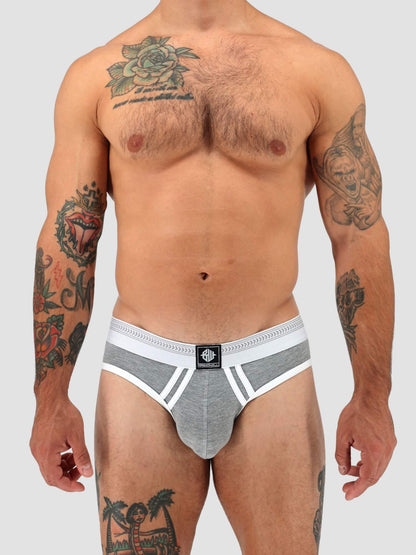 A tattooed male model wearing a gray and white low-rise brief, shown from the chest down against a solid light gray background.