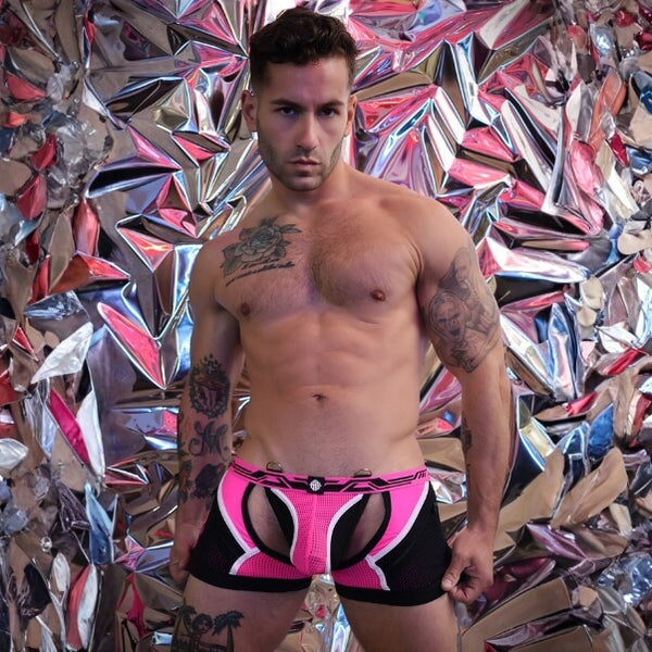 A shirtless man wearing low-rise black and hot pink mesh briefs with a front keyhole cutout, standing in front of a reflective metallic crumpled-paper wall.