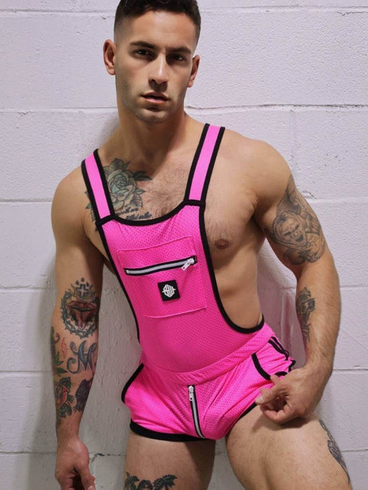 Man wearing a bright pink romper with black straps against a white brick wall.