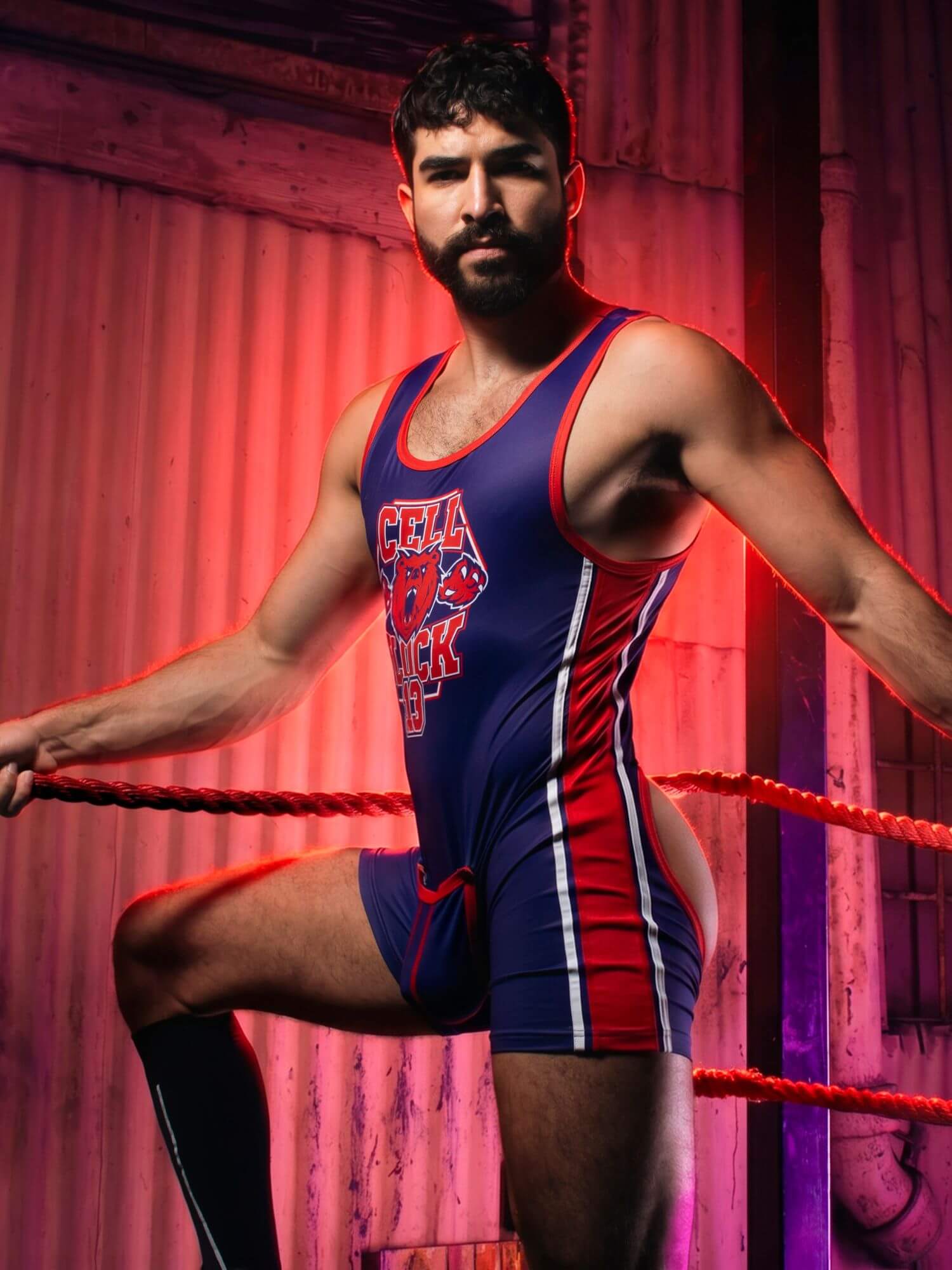 A bearded man wearing a blue, red, and white wrestling singlet stands inside a wrestling ring, posed with one leg raised. The background features a rustic, red-lit corrugated metal wall.