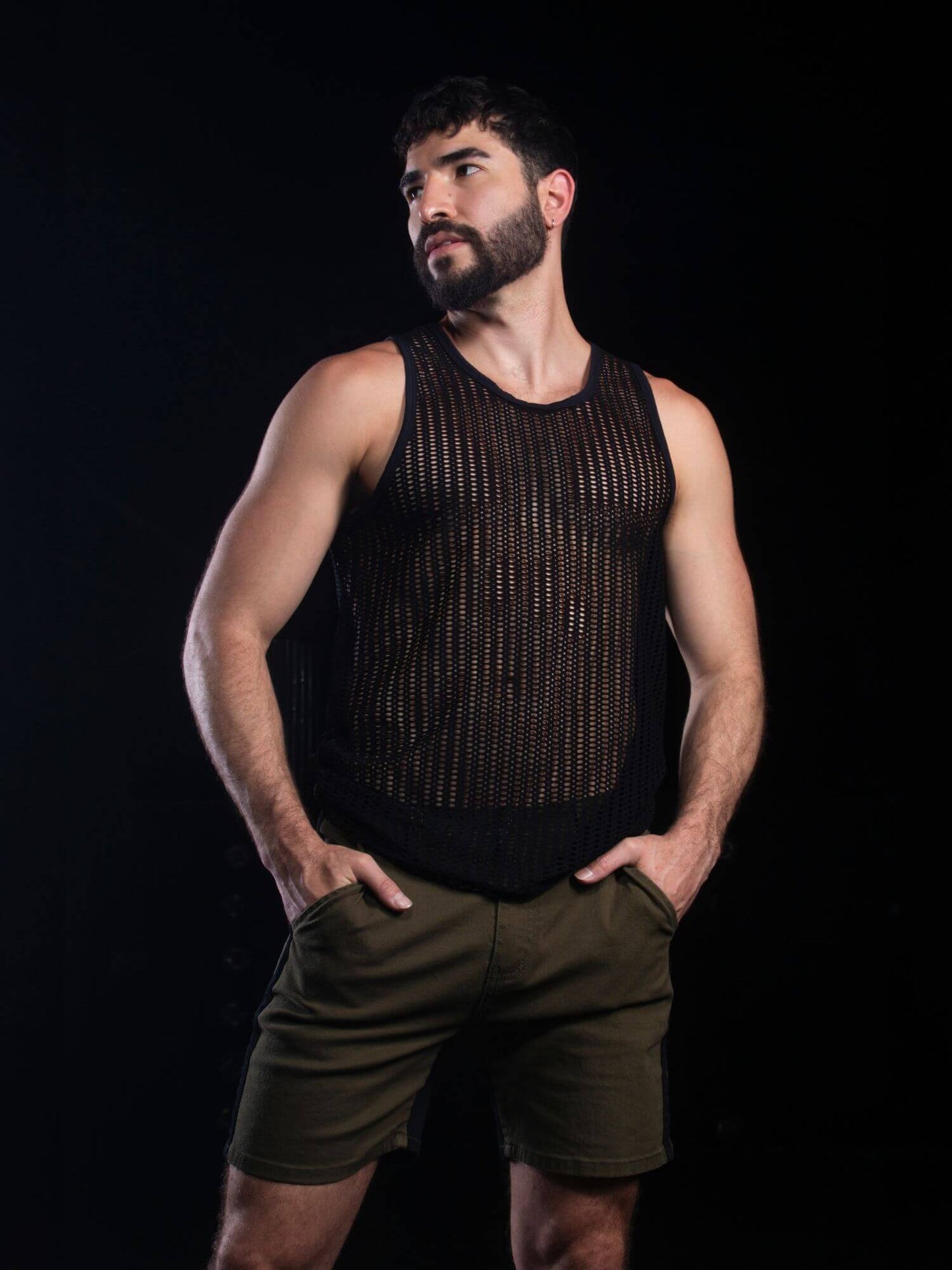 A muscular man with a beard wearing a black open-knit sleeveless tank top and olive green shorts, posing against a solid black background.