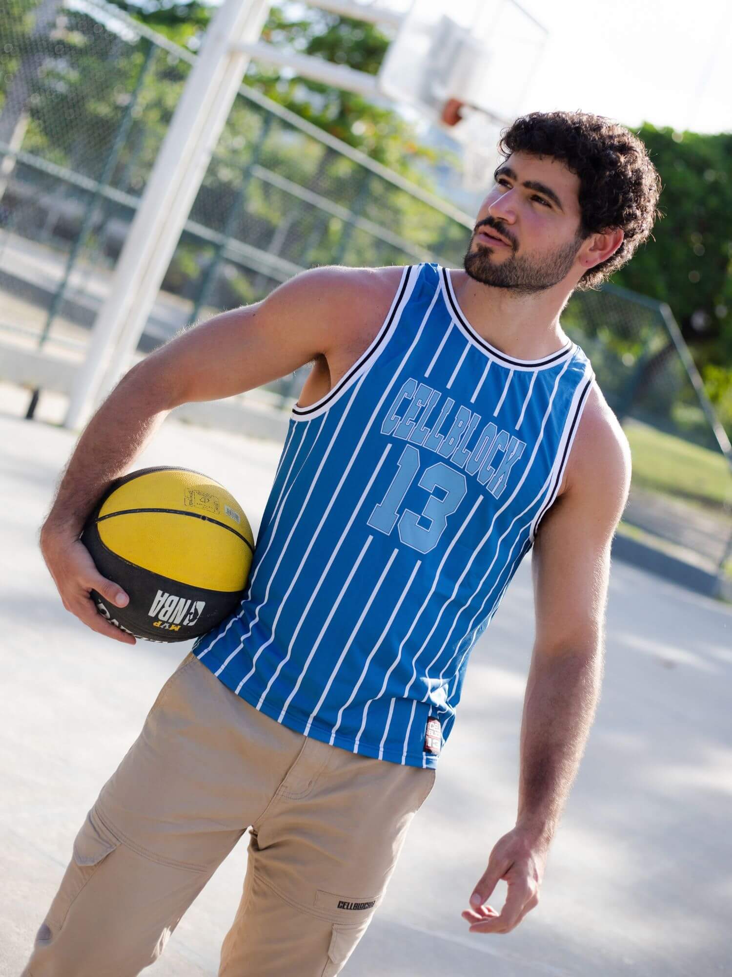 A man on an outdoor basketball court wearing a blue and white pinstriped basketball jersey with the number 13 and holding a yellow and black basketball.