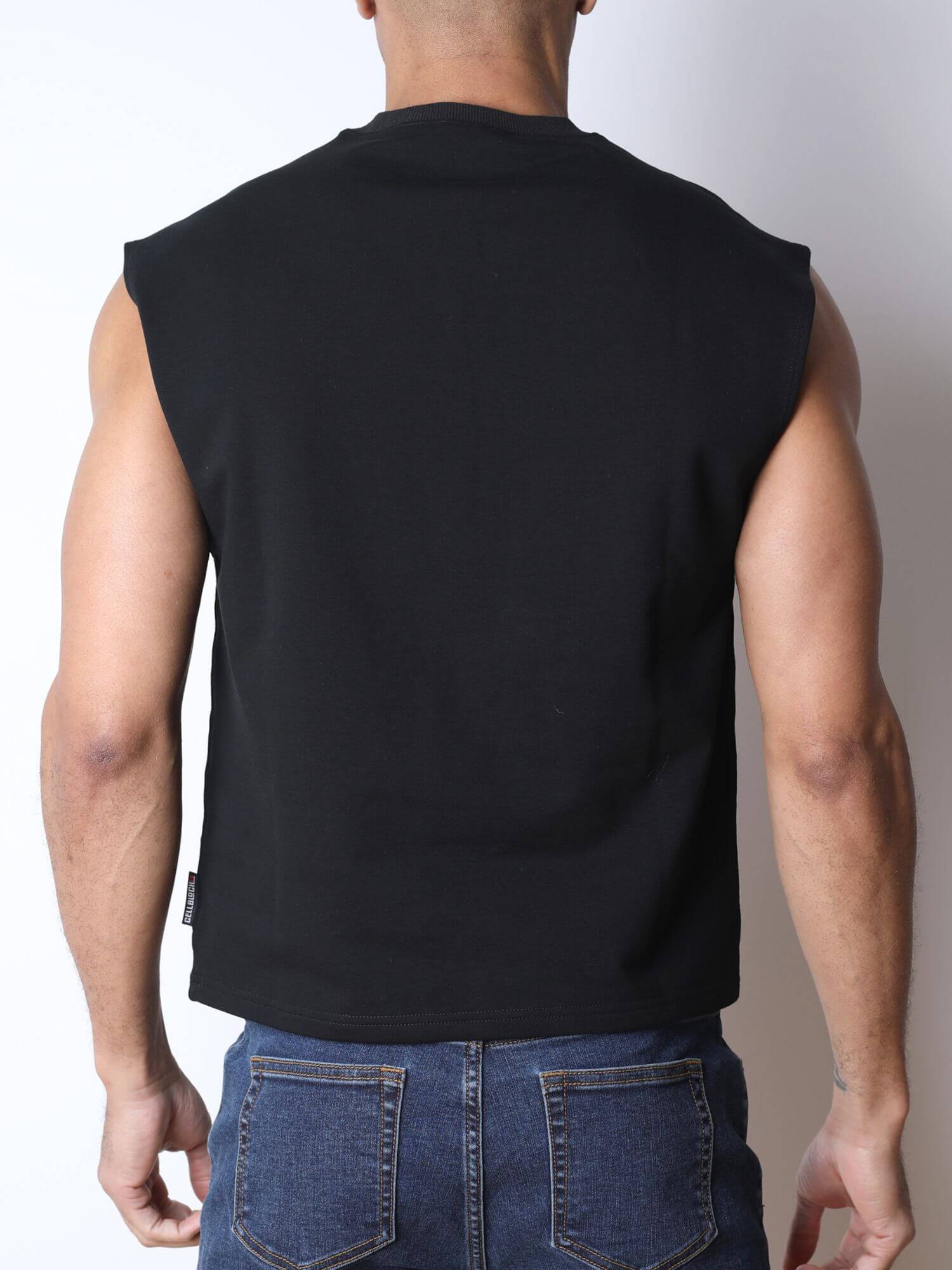 Rear view of a man wearing a black sleeveless muscle shirt and blue jeans against a plain light gray background.