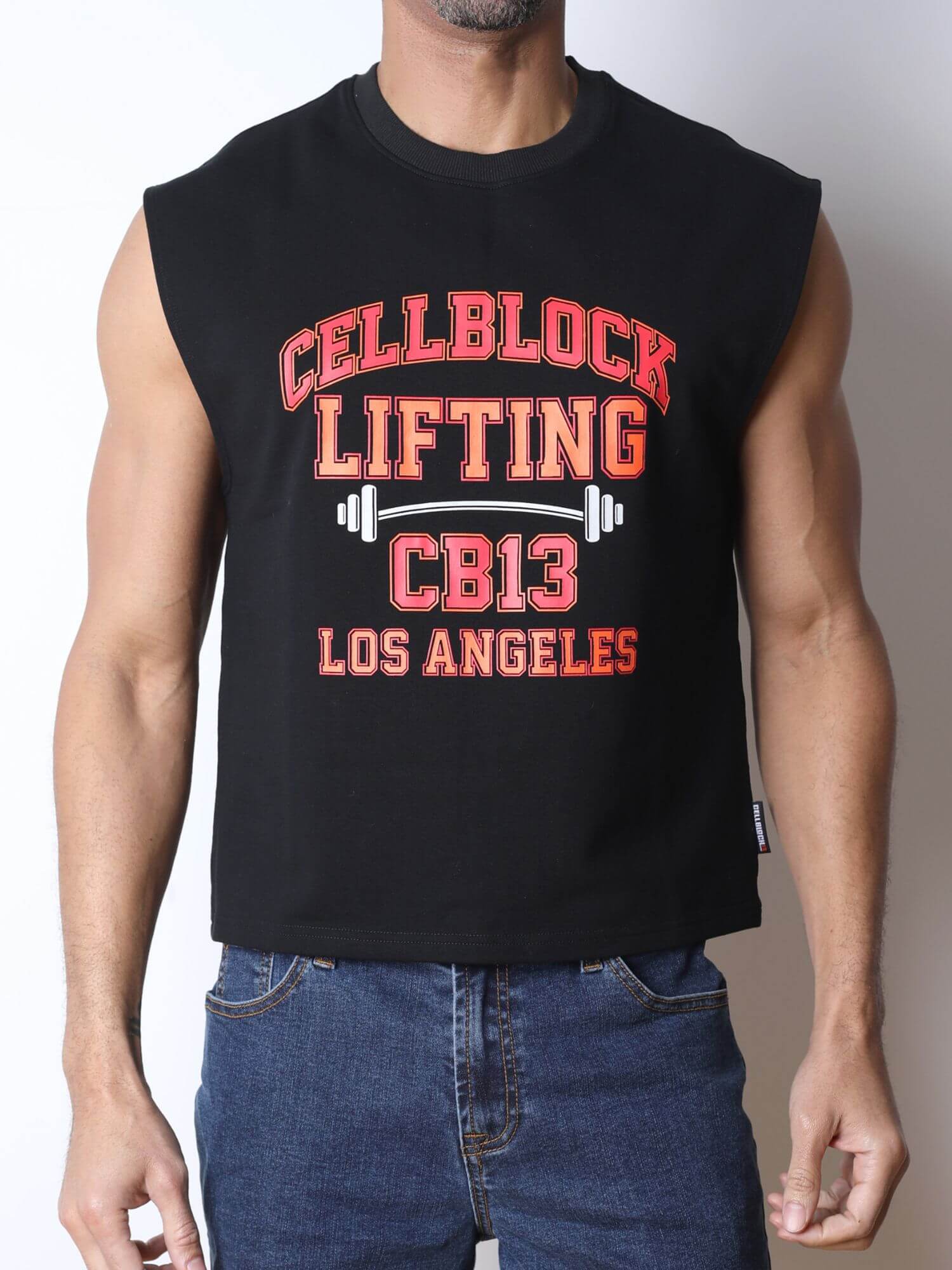 A man wearing a black sleeveless muscle tank top with a distressed red and white graphic that reads 'CELLBLOCK LIFTING CB13 LOS ANGELES' with a barbell design, paired with blue jeans against a plain light background.