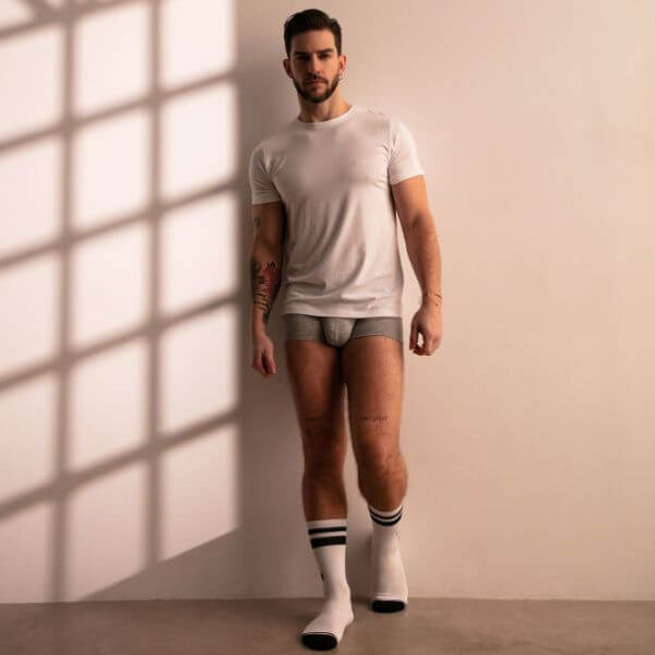 Man wearing a white t-shirt and gray underwear standing against a beige wall with sunlight casting shadows.