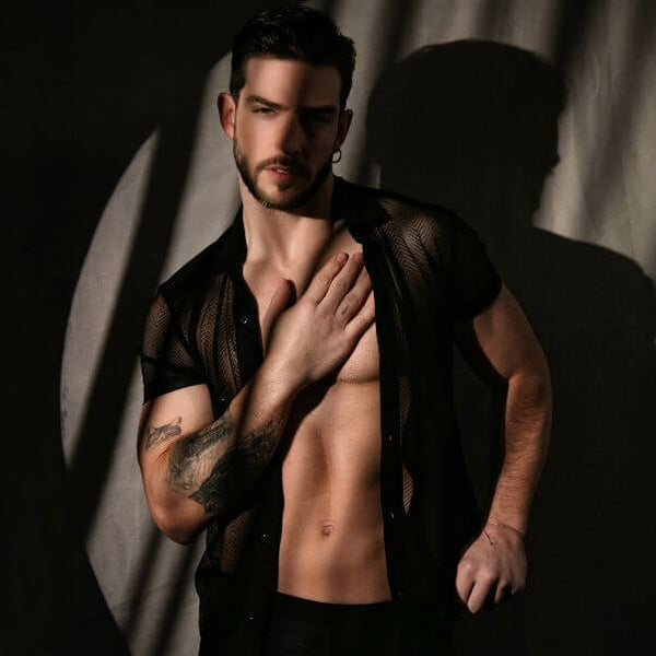 Man with a tattoo on his arm wearing a mesh shirt against a dark background