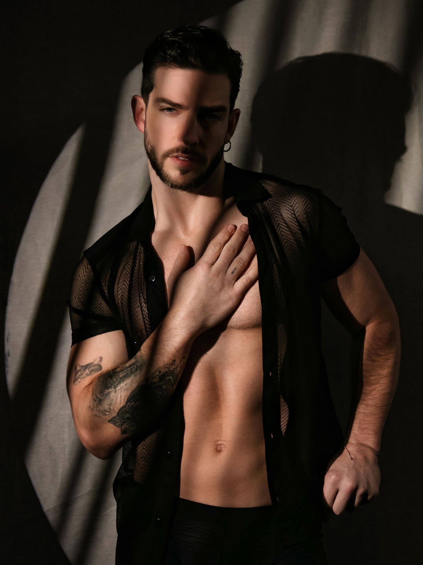 Man with tattoos wearing a sheer black shirt against a dark background