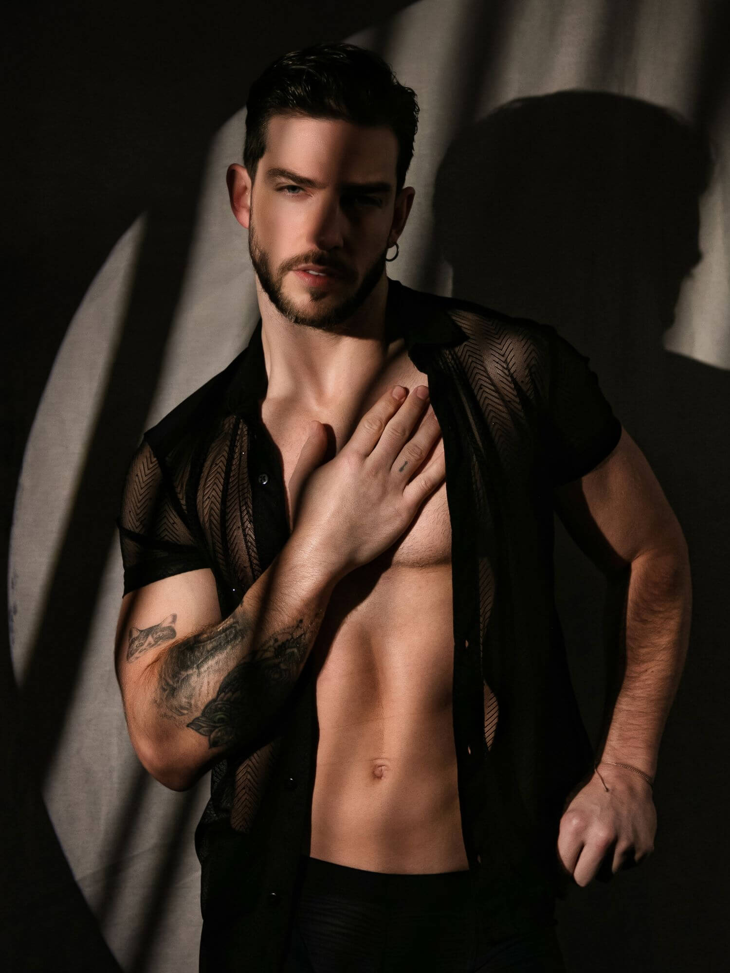 Man with tattoos wearing a sheer black shirt against a dark background