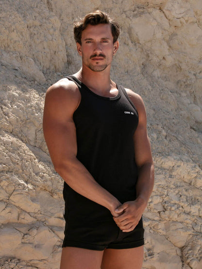 Man wearing a black tank top and shorts standing against a stone wall.