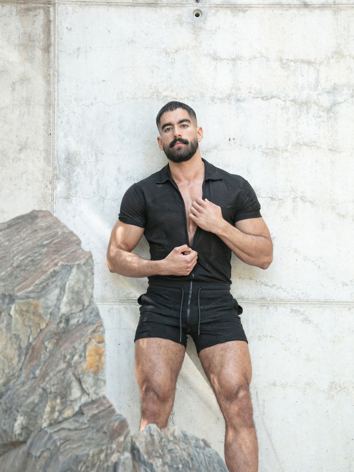 A muscular man with a beard stands against a concrete wall wearing a black short-sleeve button-up shirt and black shorts, with a large rock visible in the lower left corner.
