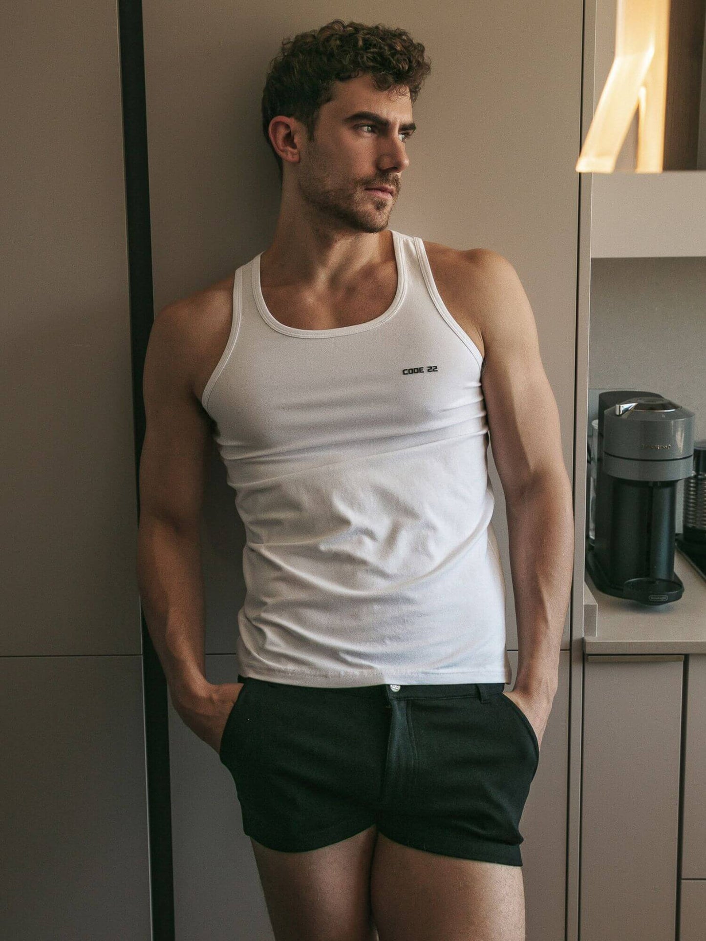 Man wearing CODE 22 white tank top and black mini shorts in a kitchen setting