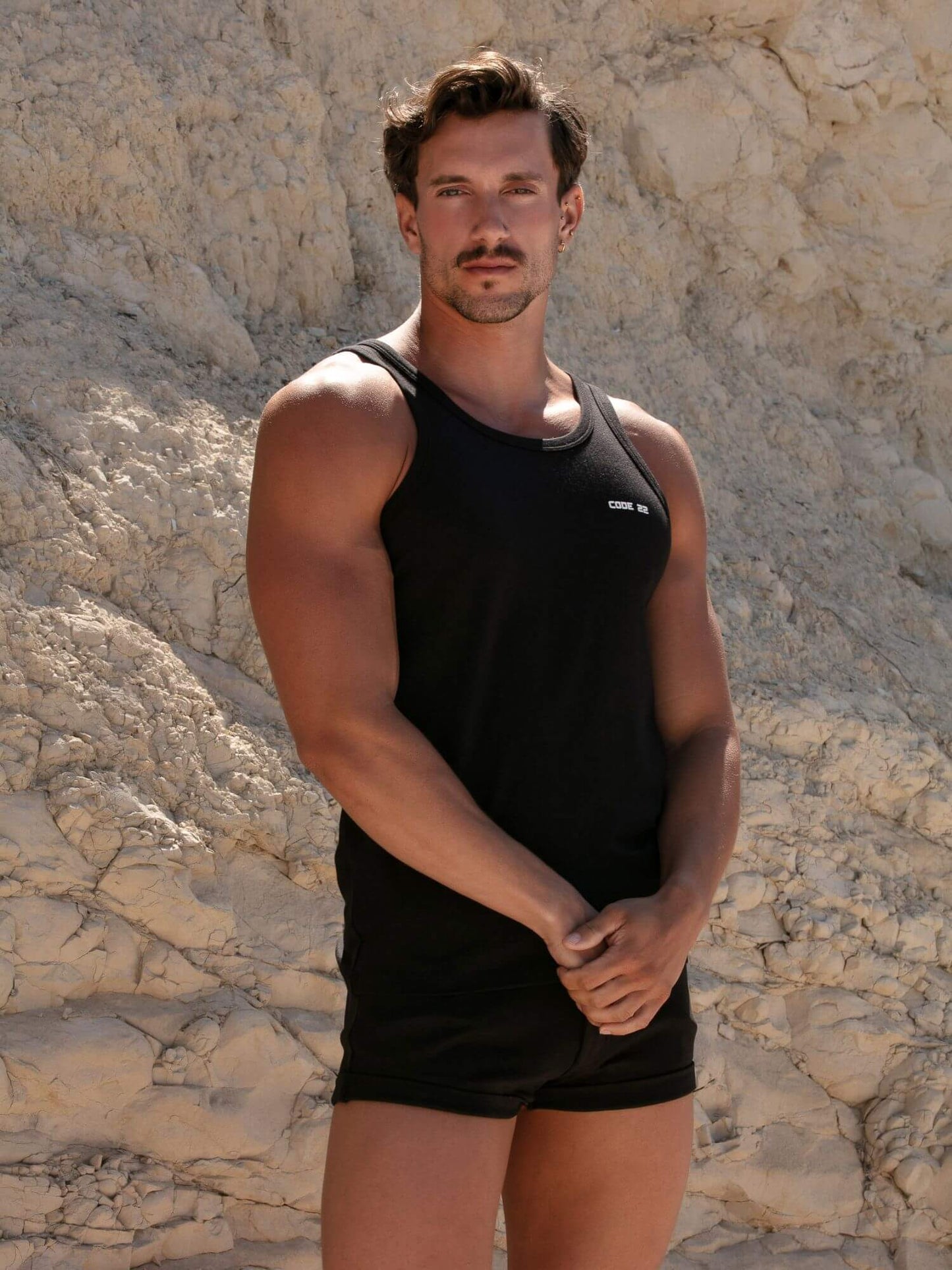 Man wearing a CODE22 black tank top and shorts standing against a stone wall.