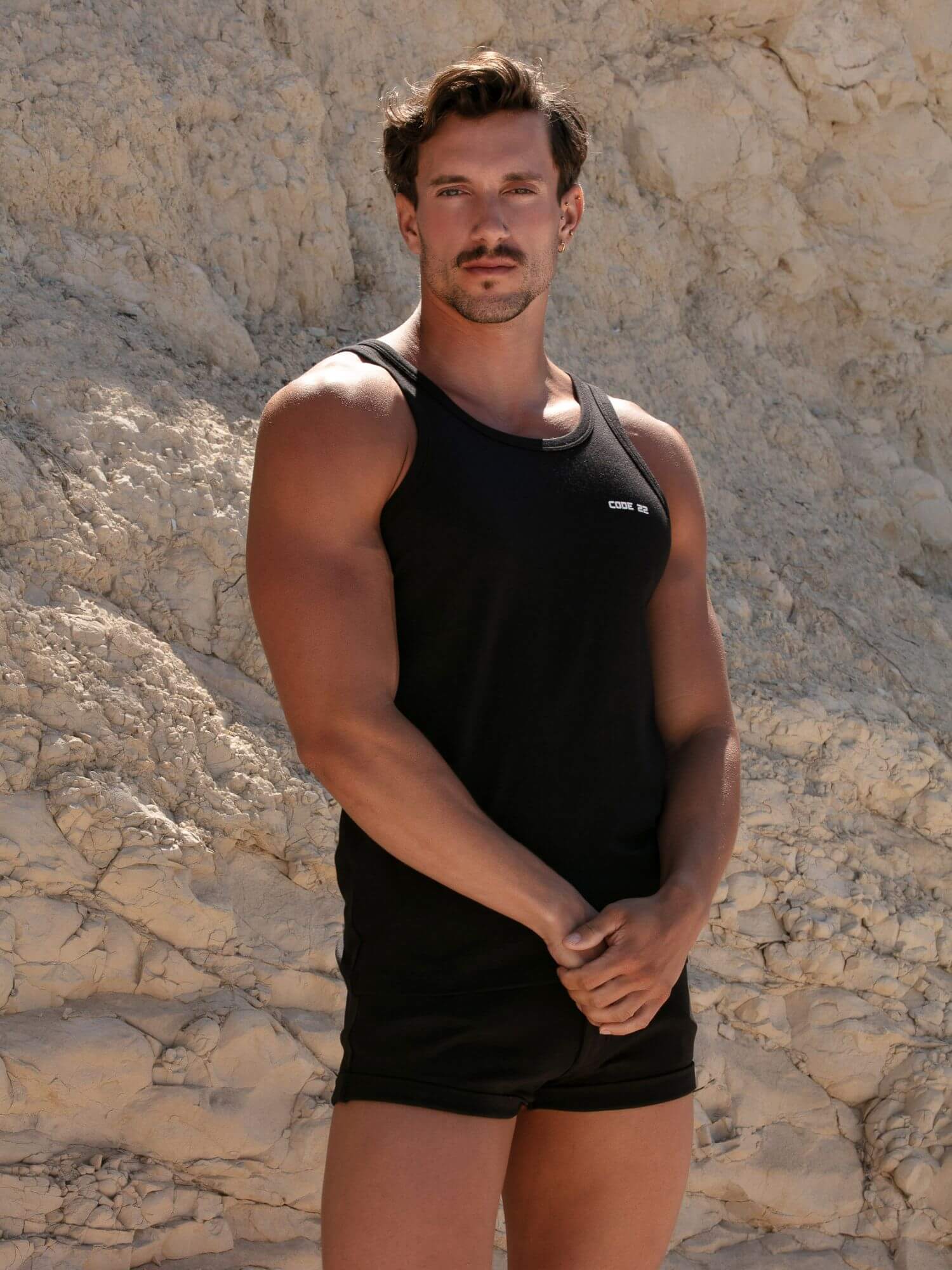 Man wearing a CODE22 black tank top and shorts standing against a stone wall.