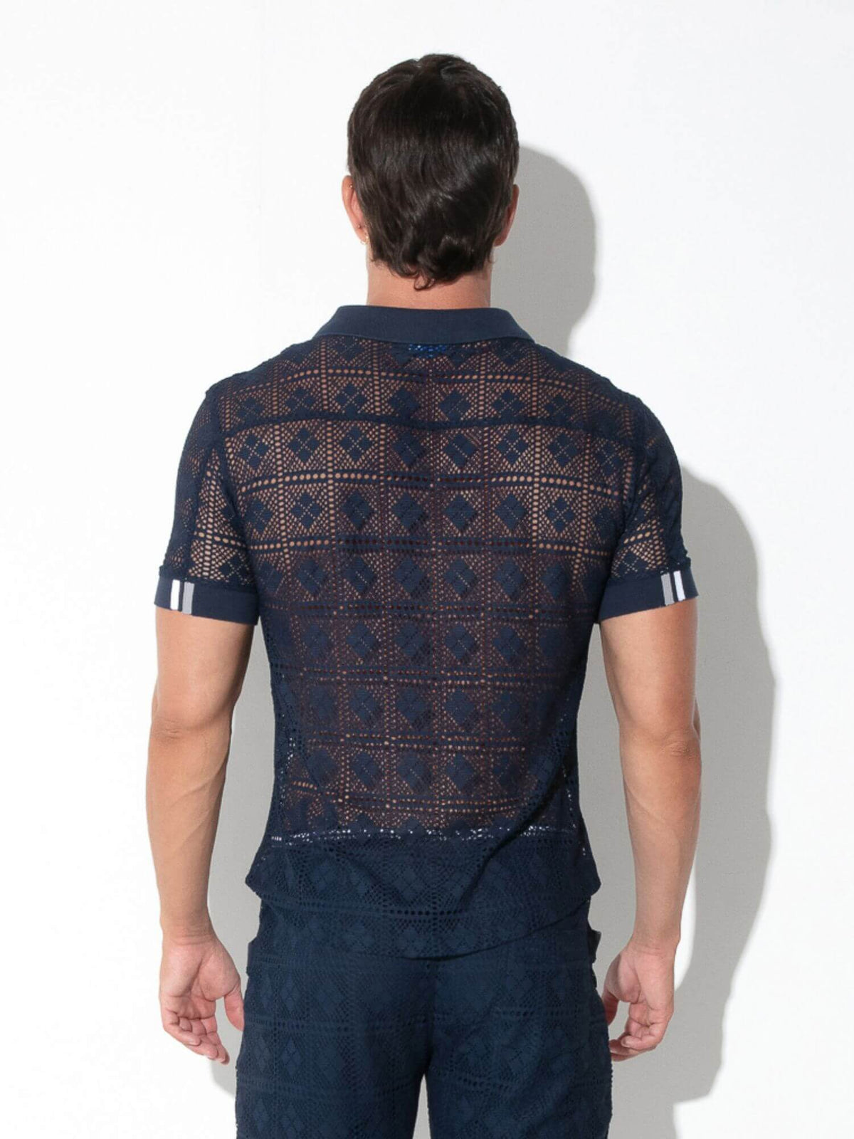A man viewed from behind wearing a navy blue short-sleeve knit polo shirt with a geometric openwork pattern and matching navy trousers, standing against a white wall.