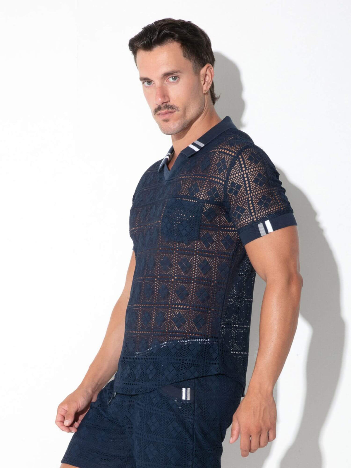 A man modeling a dark navy blue short-sleeve polo shirt with a geometric openwork pattern, paired with matching shorts, against a white background with a soft shadow.