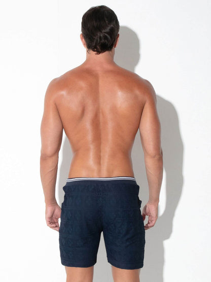 A man shown from behind wearing dark navy blue swim trunks with a white elastic waistband, standing against a white wall with a soft shadow.
