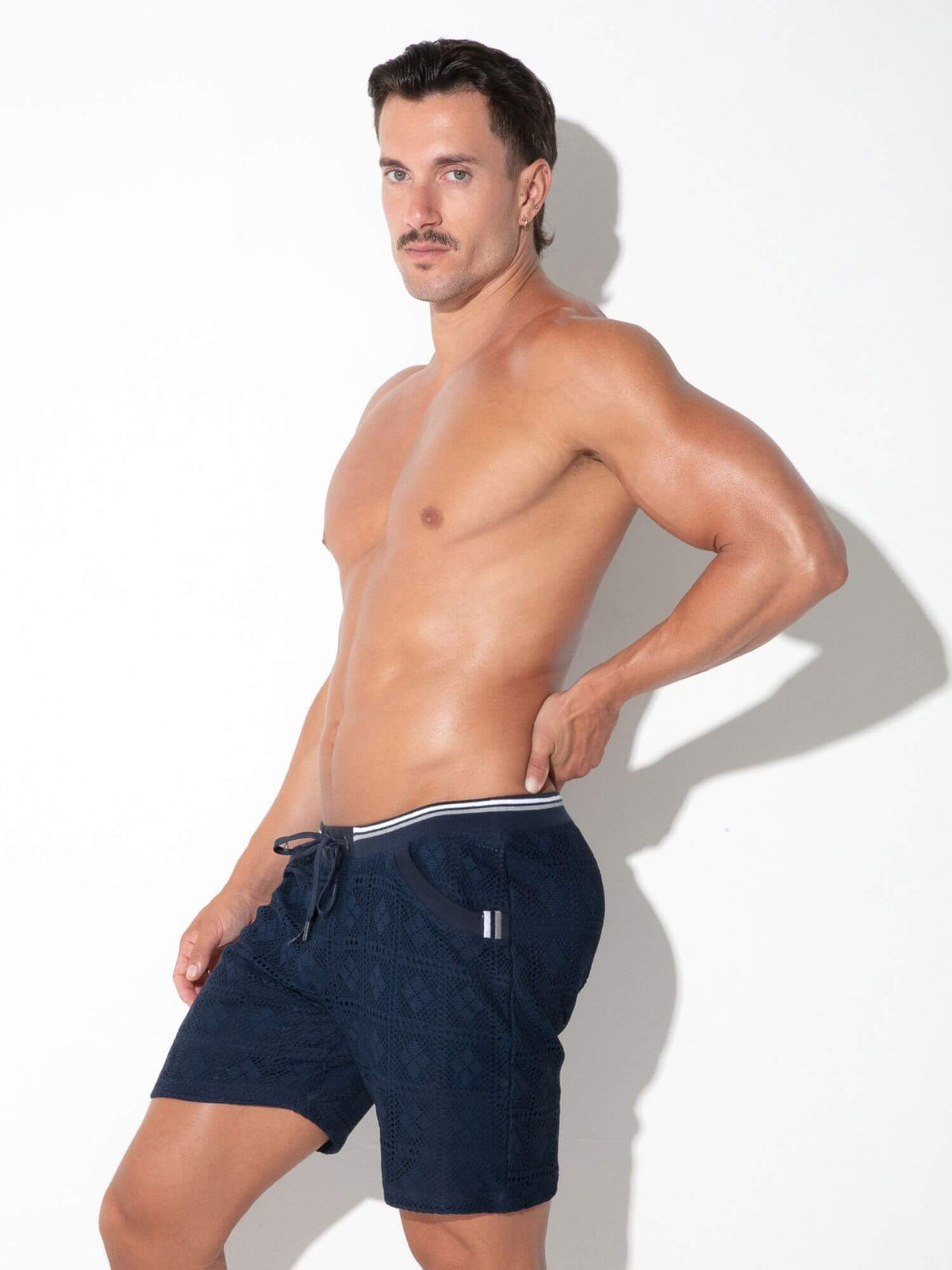 A shirtless man modeling dark navy drawstring swim shorts with a white stripe detail, posed against a white studio background.