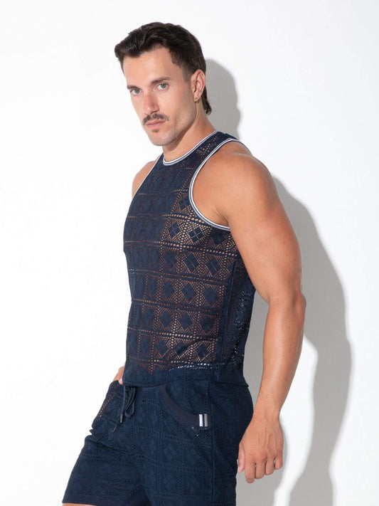 A muscular man with a mustache wearing a dark navy blue sleeveless knit vest with geometric openwork patterns and matching dark shorts, posing against a white studio background with a soft shadow.