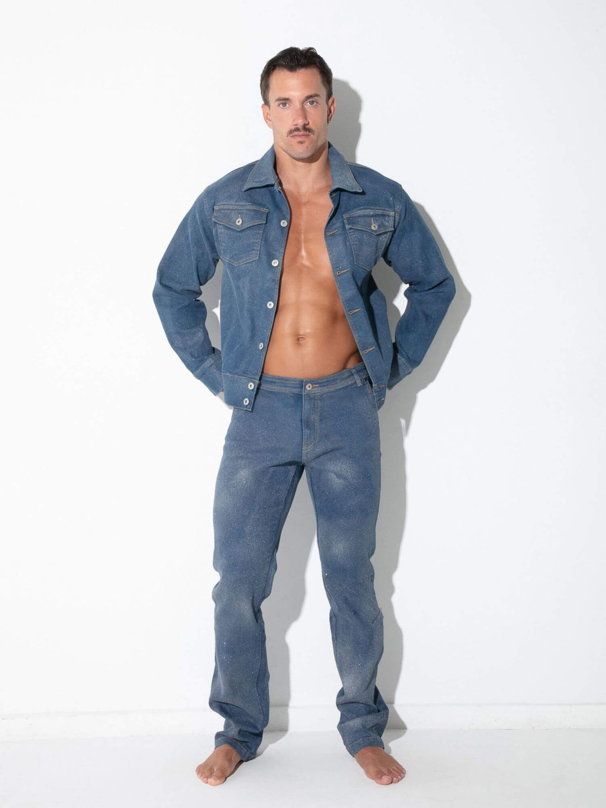 A man modeling a matching blue denim jacket and jeans set against a plain white studio background.