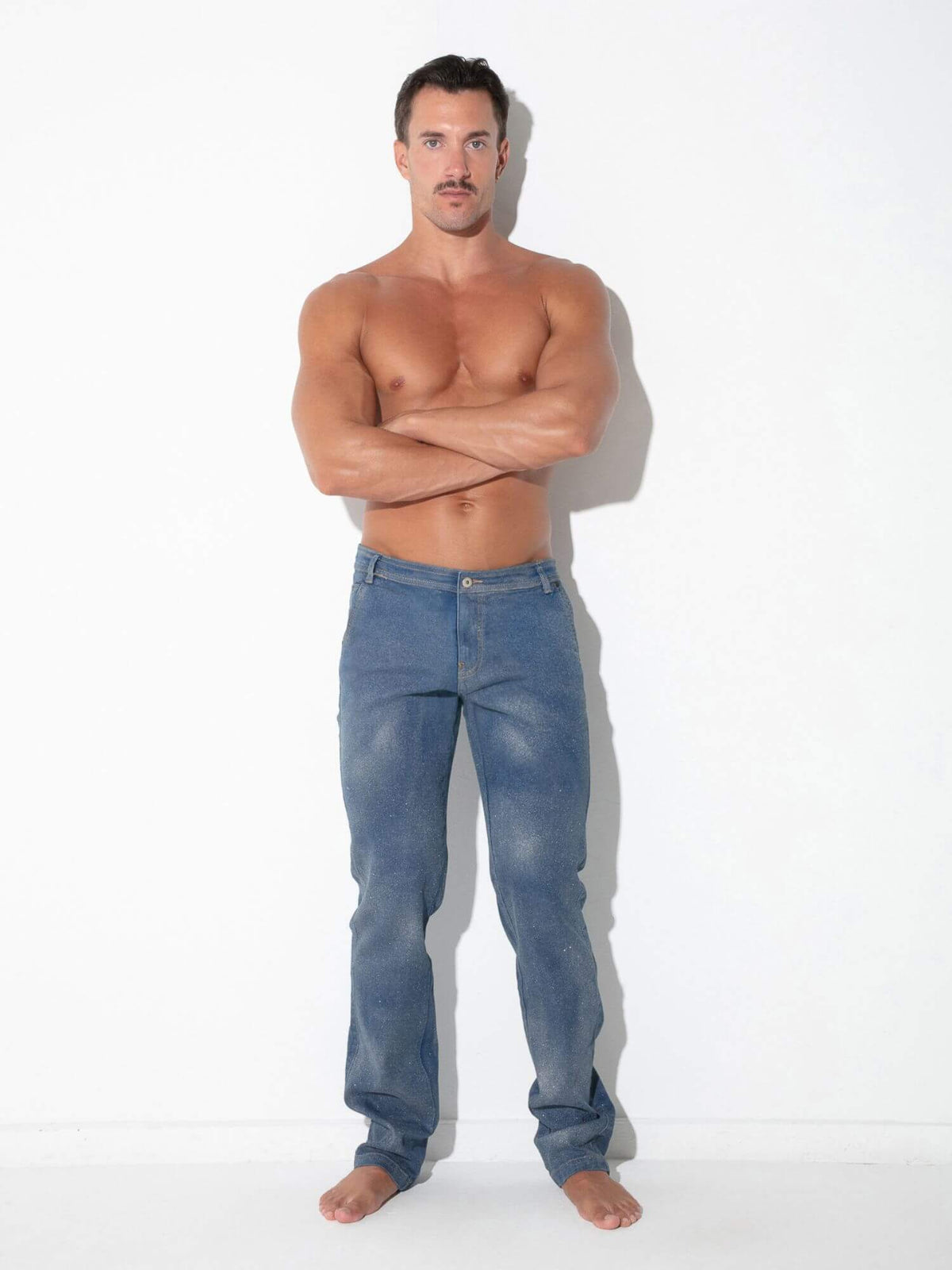 A shirtless muscular man with a mustache stands barefoot against a white wall, wearing blue jeans with his arms crossed.