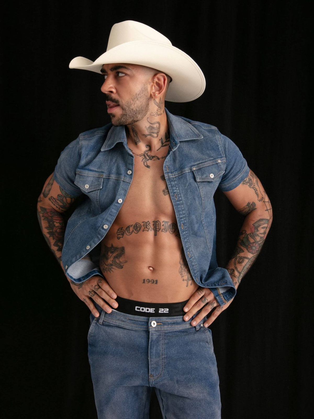 A man wearing a white cowboy hat, an open blue denim shirt, and blue jeans with a visible Code 22 waistband, posed against a black background.